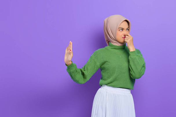 Beautiful annoyed Asian woman in green sweater makes stop gesture, covering her nose with hand isolated over purple background - Fotografie, Obrázek