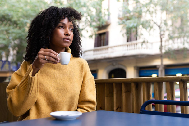 Молодая афроамериканка держит чашку кофе на открытой террасе кафе. Копирование пространства. - Фото, изображение