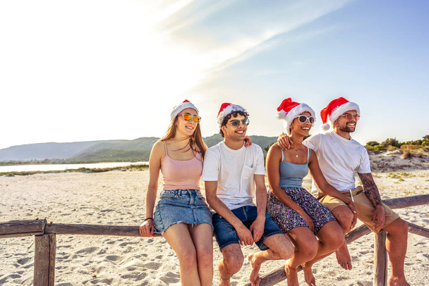 Happy people enjoying outdoor life in exotic vacation resort sitting on wooden fence on a white sand beach at sunset. Group of young friends having fun together Santa Claus hat at sea vacation resort - Photo, Image