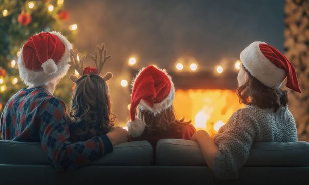 メリークリスマスとハッピーホリデー。ママとパパと可愛い娘たちが暖炉を見てる。室内で木のそばで遊ぶ親子. - 写真・画像
