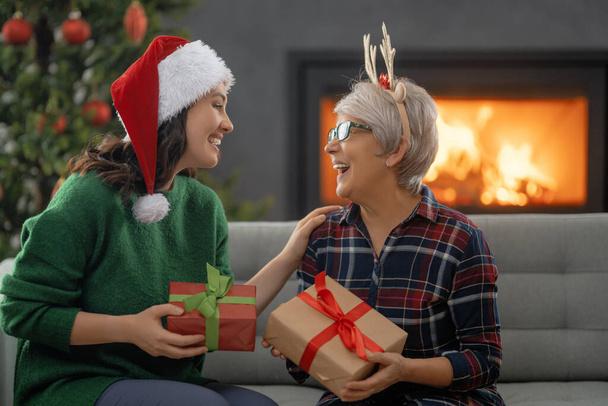 Merry Christmas and Happy Holidays. Senior mom and her adult daughter exchanging gifts. Having fun near tree indoors. Loving family with presents in room.  - Фото, изображение