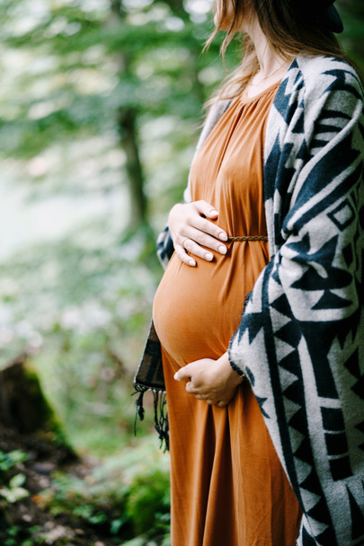 Η έγκυος αγκαλιάζει την κοιλιά της με τα χέρια της στο πάρκο.  - Φωτογραφία, εικόνα
