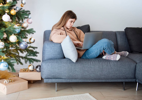 Девочка-подросток с ноутбуком сидит на диване в гостиной рядом с елкой - Фото, изображение