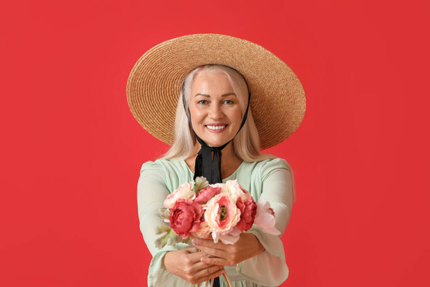 Mature woman in straw hat holding bouquet of beautiful flowers on red background - Photo, Image