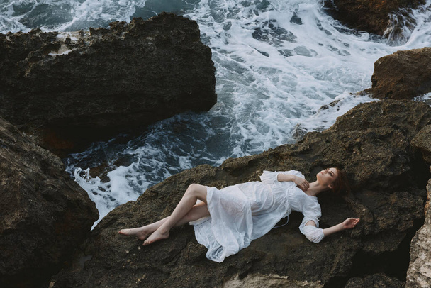 atraktivní žena s dlouhými vlasy v dlouhých bílých šatech mokré vlasy ležící na skalnatém útesu pohled shora - Fotografie, Obrázek