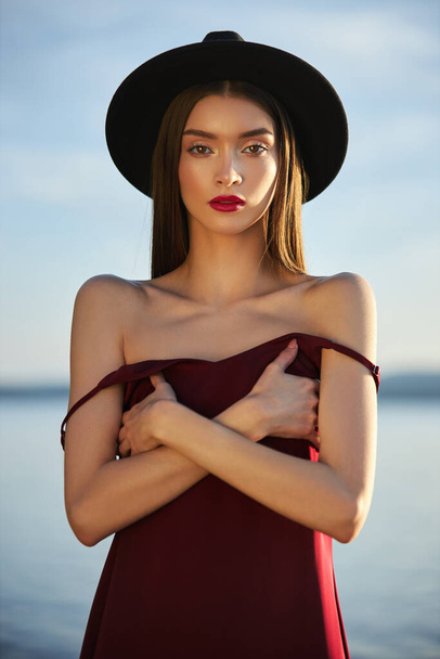 Portrait of a beautiful woman with long hair in a big round black hat. Beauty skin face, bare shoulders. Girl golden light at sunset - Photo, image