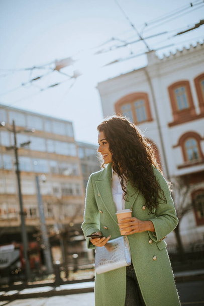 Csinos fiatal nő használja okostelefon az utcán, és tartja elvitelre kávé - Fotó, kép