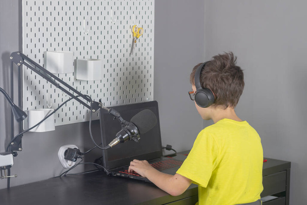 A teenage boy in a yellow T-shirt is blogging at home in front of a laptop with a microphone and headphones. - Foto, imagen