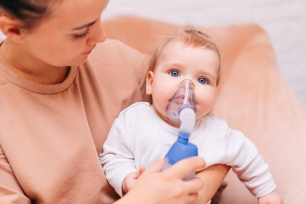 Pieni tyttö hengittää inhalaattorista, jossa on sumutin kotona. Lapsi, jolla on virustauti hengityssuojaimessa, istuu äitinsä käsivarsissa. Astmaa sairastava potilas - Valokuva, kuva