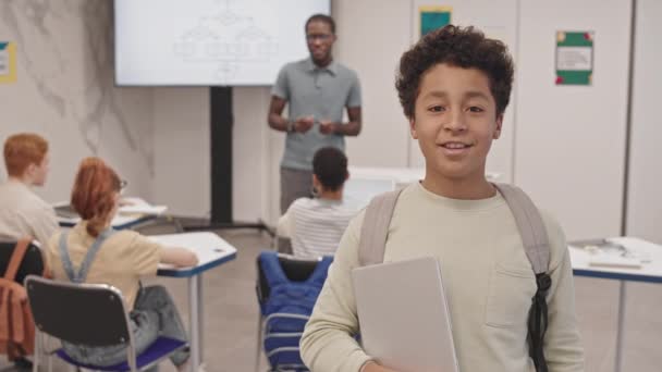 Наклонение веселого Бирасиал мальчика в рюкзаке, держа ноутбук, стоя в классе во время урока, разговаривая, улыбаясь и глядя в камеру - Кадры, видео