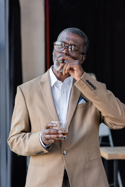 Senior afrikanisch-amerikanischer Geschäftsmann raucht Zigarre, während er ein Glas Whiskey in der Hand hält - Foto, Bild