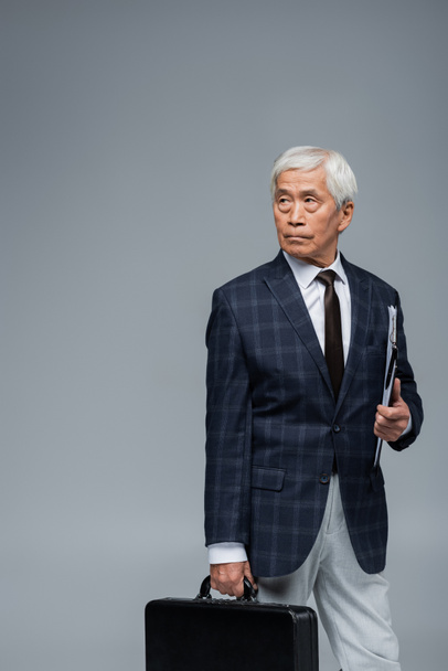serio asiático hombre de negocios con maletín y portapapeles mirando lejos aislado en gris - Foto, imagen