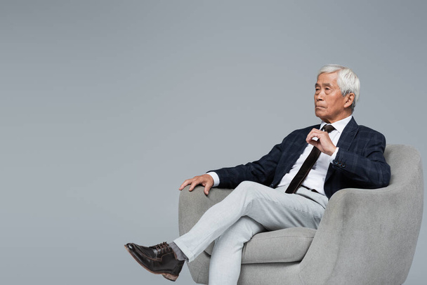 senior y confidente asiático hombre de negocios mirando hacia otro lado mientras sentado en sillón aislado en gris - Foto, imagen
