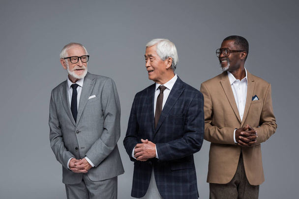 elderly multiethnic businessmen in formal wear smiling at each other isolated on grey - Foto, Imagem