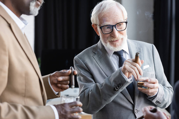 senior businessman with cigar and whiskey looking away during discussion with blurred african american colleague - Zdjęcie, obraz