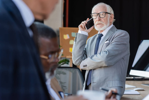 літній бізнесмен в окулярах розмовляє на мобільному телефоні біля розмитих багатоетнічних колег
 - Фото, зображення