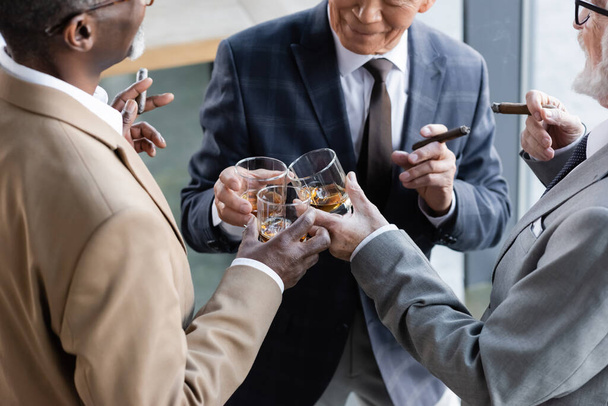 bijgesneden beeld van senior succesvolle zakenpartners met sigaren klinkende glazen whisky op kantoor - Foto, afbeelding