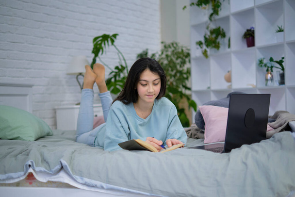 Teenage girl doing homework on the bed. teen girl school student write notes watch video online webinar learn on laptop. distance learning course video conference pc call. - Photo, Image