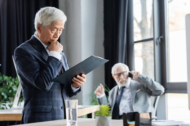 thoughtful asian businessman looking at clipboard near senior colleague talking on smartphone on blurred background - Фото, зображення