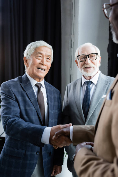 vrolijk aziatische zakenman schudden handen met wazig Afrikaans amerikaanse man in de buurt van senior collega - Foto, afbeelding
