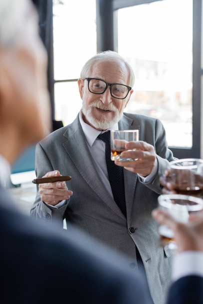 hombre de negocios senior con cigarro y vaso de whisky hablando con un colega borroso en la oficina - Foto, Imagen
