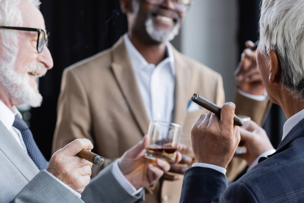 senior businessman holding glass of whiskey and cigar near smiling multiethnic colleagues - Фото, зображення