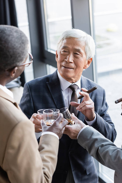 senior asian businessman smiling while clinking glasses of whiskey with business partners - Фото, изображение