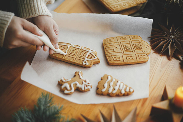 Hände dekorieren weihnachtliche Lebkuchen mit Zuckerguss auf einem Holztisch mit Kerzen und Ornamenten. Nahaufnahme der Herstellung von Lebkuchenhaus mit Zuckerguss. Stimmungsvolles Stimmungsbild. Urlaubsvorbereitungen - Foto, Bild