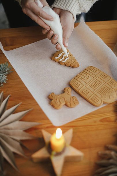 Mani che decorano i biscotti di pan di zenzero di Natale con ciliegina sul tavolo di legno con candela e ornamenti. Chiudete. Fare casa di pan di zenzero con glassa. Preparazione e tradizione delle vacanze - Foto, immagini