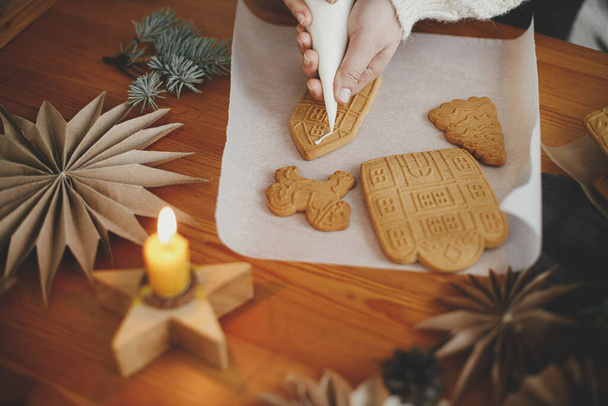 Ręce zdobiące świąteczne ciasteczka piernikowe z lukrem na drewnianym stole ze świecami i ozdobami. Zamknij drzwi. Robię domek z piernika z lukrem. Przygotowanie wakacji i tradycja - Zdjęcie, obraz