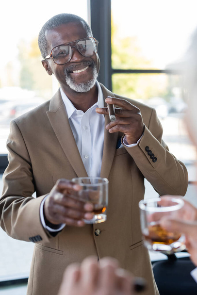 senior african american businessman with cigar and glass of whiskey smiling near blurred colleague in office - Photo, Image