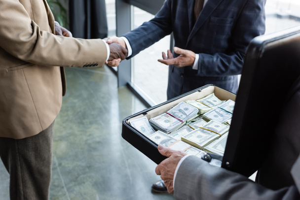 vista parcial de colegas de negocios interracial estrechando la mano cerca de un hombre sosteniendo el maletín con dinero - Foto, imagen