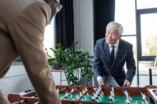 borrosa africano americano hombre de negocios jugando fútbol de mesa con senior asiático colega - Foto, Imagen