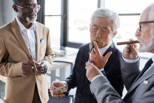 Afrika kökenli Amerikalı iş ortağının yanında Asyalı meslektaşının purosunu yakan kıdemli iş adamı. - Fotoğraf, Görsel