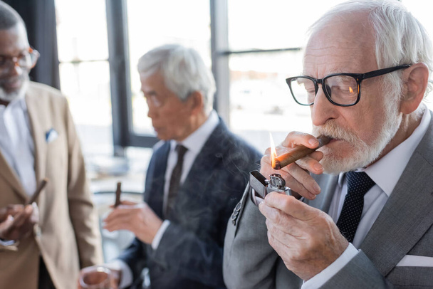 senior successful businessman lighting cigar near blurred multiethnic business partners talking in office - 写真・画像
