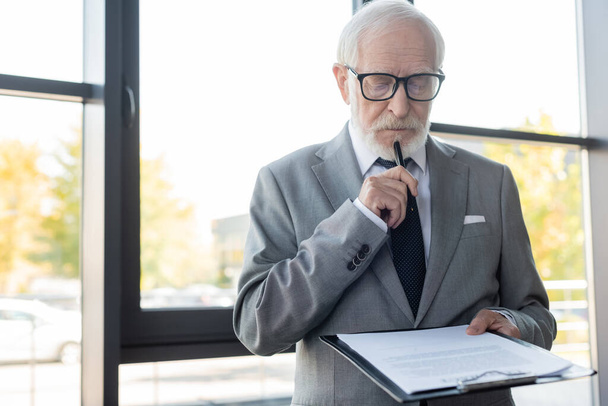 thoughtful senior businessman holding pen while reading contract in office - 写真・画像