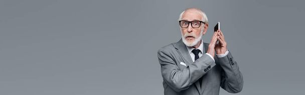 shocked senior businessman looking at camera while covering cellphone with hand isolated on grey, banner - Фото, изображение