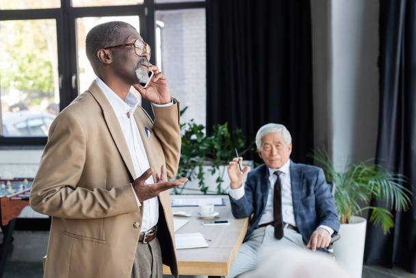 african american businessman talking on mobile phone near blurred asian colleague - Фото, изображение
