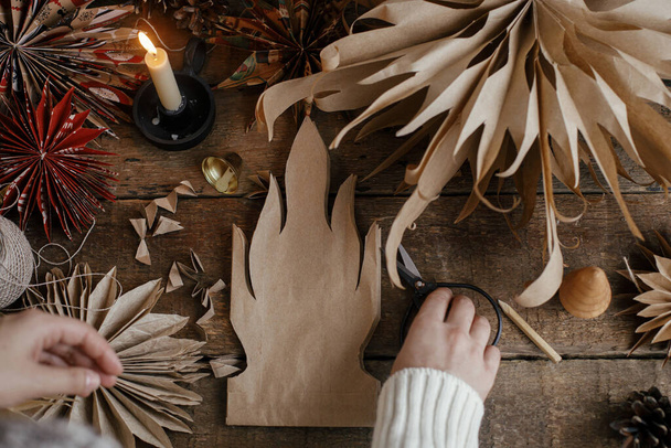 Stijlvolle kerststerren maken. Handen snijden ster van ambachtelijke papieren zak op de achtergrond van handgemaakte Zweedse sterren, draad, kaars, schaar op rustiek hout. Top view proces van het maken van feestelijke decor - Foto, afbeelding