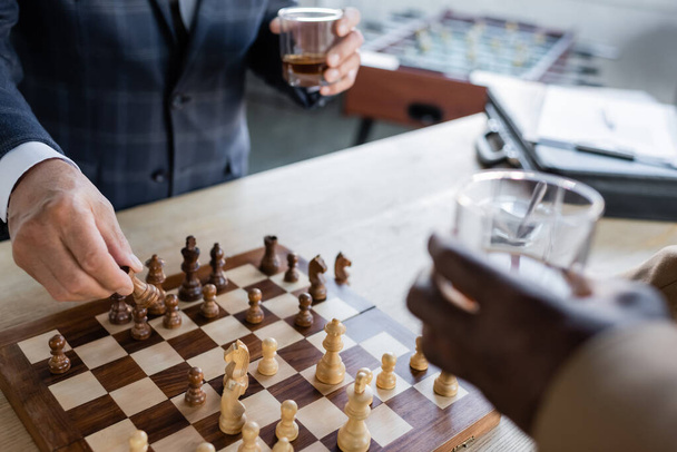 partial view of blurred senior businessmen with glasses of whiskey playing chess in office - Foto, Imagen