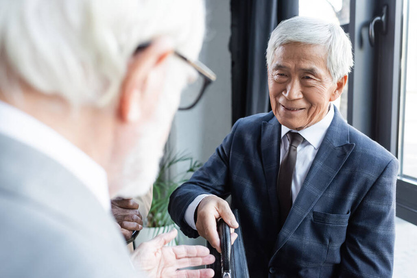 sênior asiático empresário sorrindo enquanto dando pasta para desfocado parceiro de negócios - Foto, Imagem