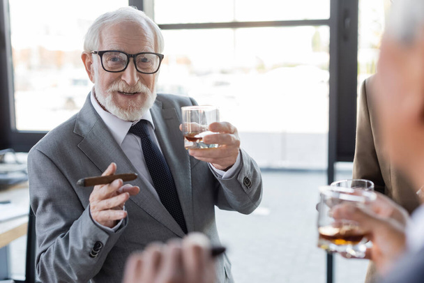 senior businessman with glass of whiskey and cigar near blurred colleagues in office - 写真・画像