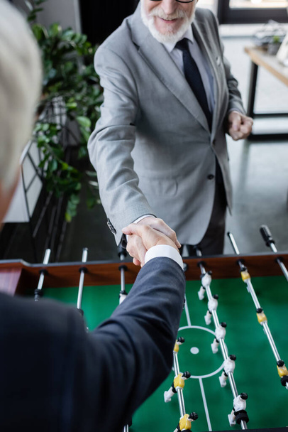 cropped view of senior businessman shaking hands with blurred colleague near table football - Foto, imagen