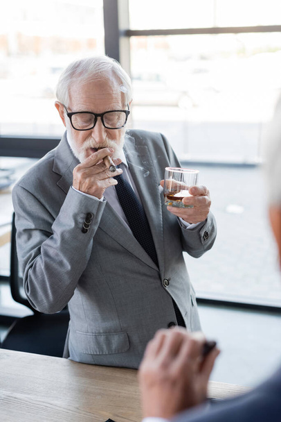 elderly businessman in eyeglasses holding glass of whiskey while smoking cigar near blurred colleague - Photo, Image