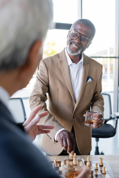 cheerful african american businessman with glass of whiskey playing chess with blurred colleague in office - Photo, Image