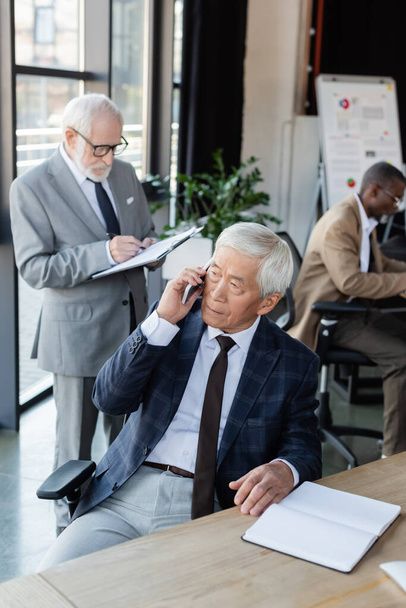 senior asiático hombre de negocios hablando en smartphone cerca vacío notebook y interracial colegas - Foto, imagen