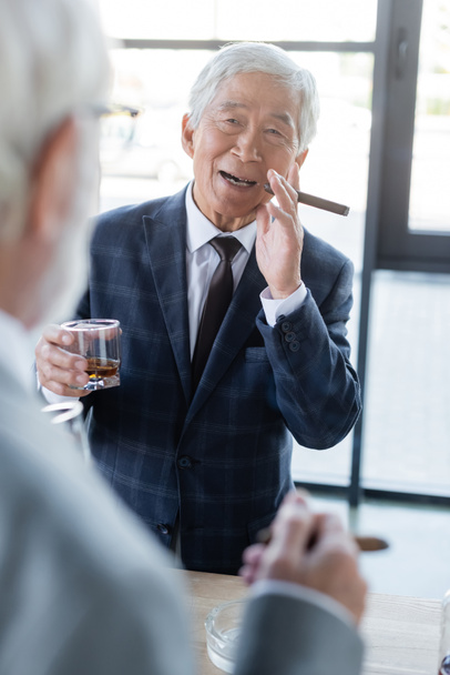 elderly asian businessman with cigar and glass of whiskey smiling near blurred colleague  - Fotó, kép
