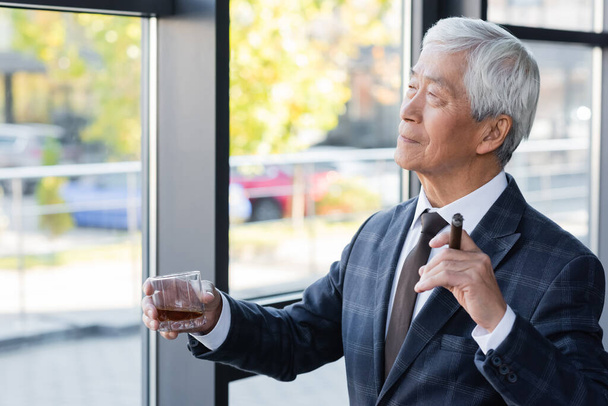 anciano asiático hombre de negocios mirando hacia otro lado mientras de pie cerca de ventana con cigarro y vaso de whisky - Foto, Imagen