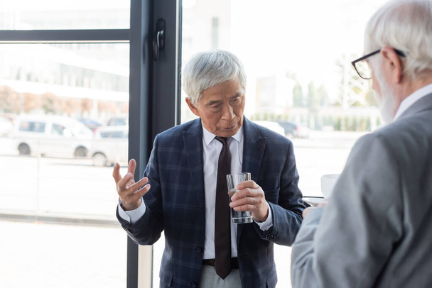 Старший азійський бізнесмен тримає склянку води під час обговорення з розмитим колегою. - Фото, зображення