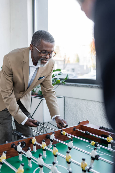 vanhempi afrikkalainen amerikkalainen liikemies pelaa pöytäjalkapalloa hämärtyneen kollegansa kanssa - Valokuva, kuva